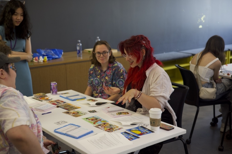 Group of Tisch Summer High school Game Design students sit at a table looking at colorful photos and notebooks during a game design class.